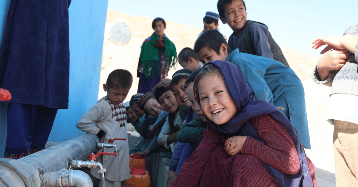 Children are sitting next to a water pipe, with a little girl in the foreground laughing